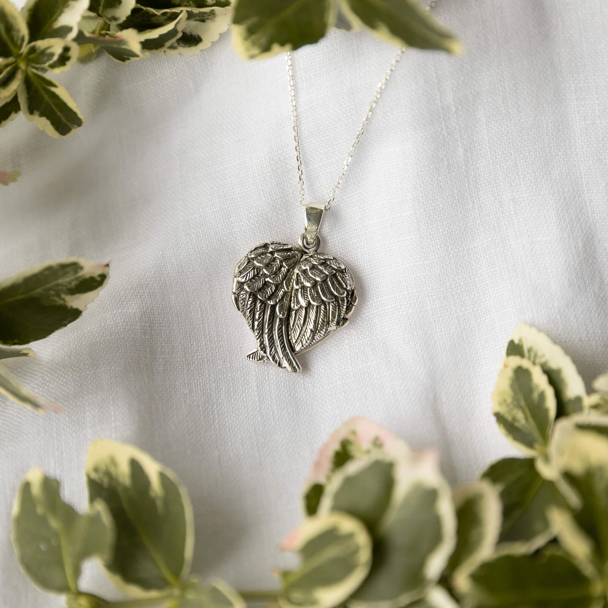 Silver angel wing necklace on a white fabric background with green leaves.