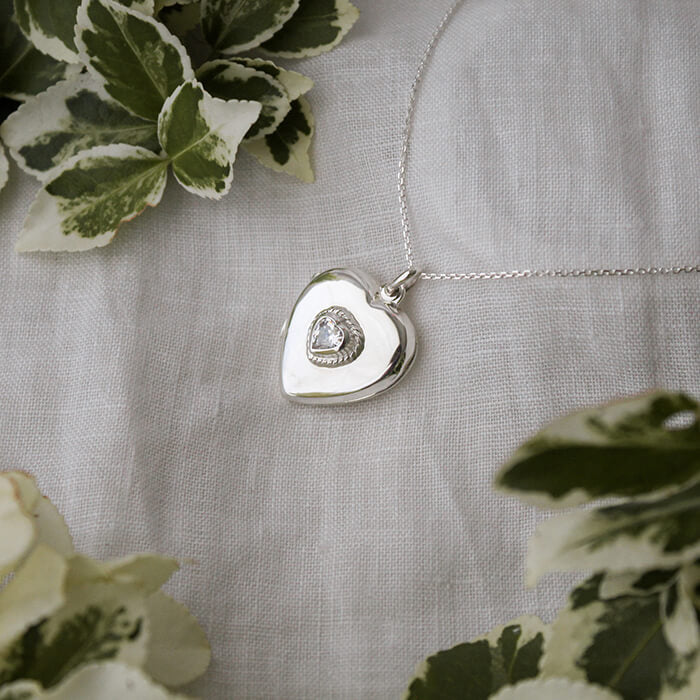 Heart shaped sterling silver locket with cubic zirconia on a linen background with some green vegetation. 