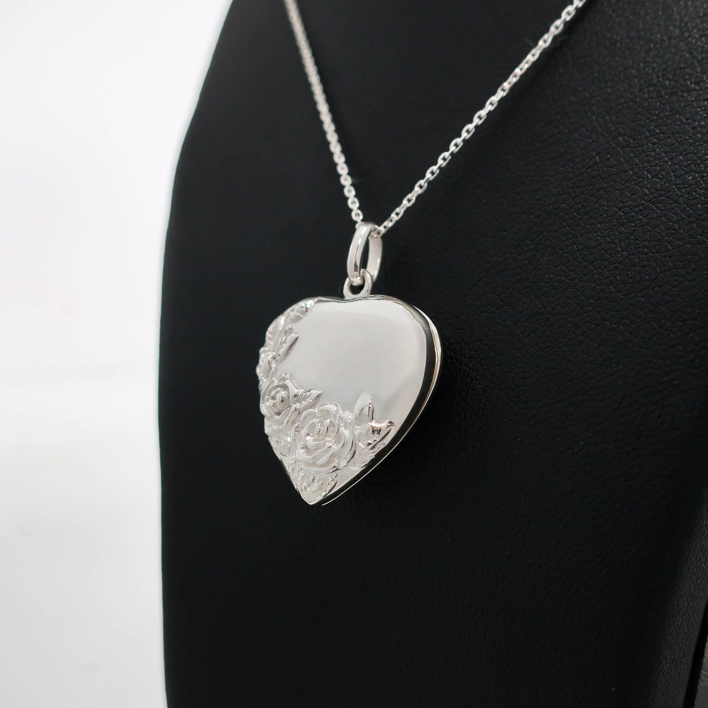 Silver heart-shaped locket with floral engraving on a black stand.